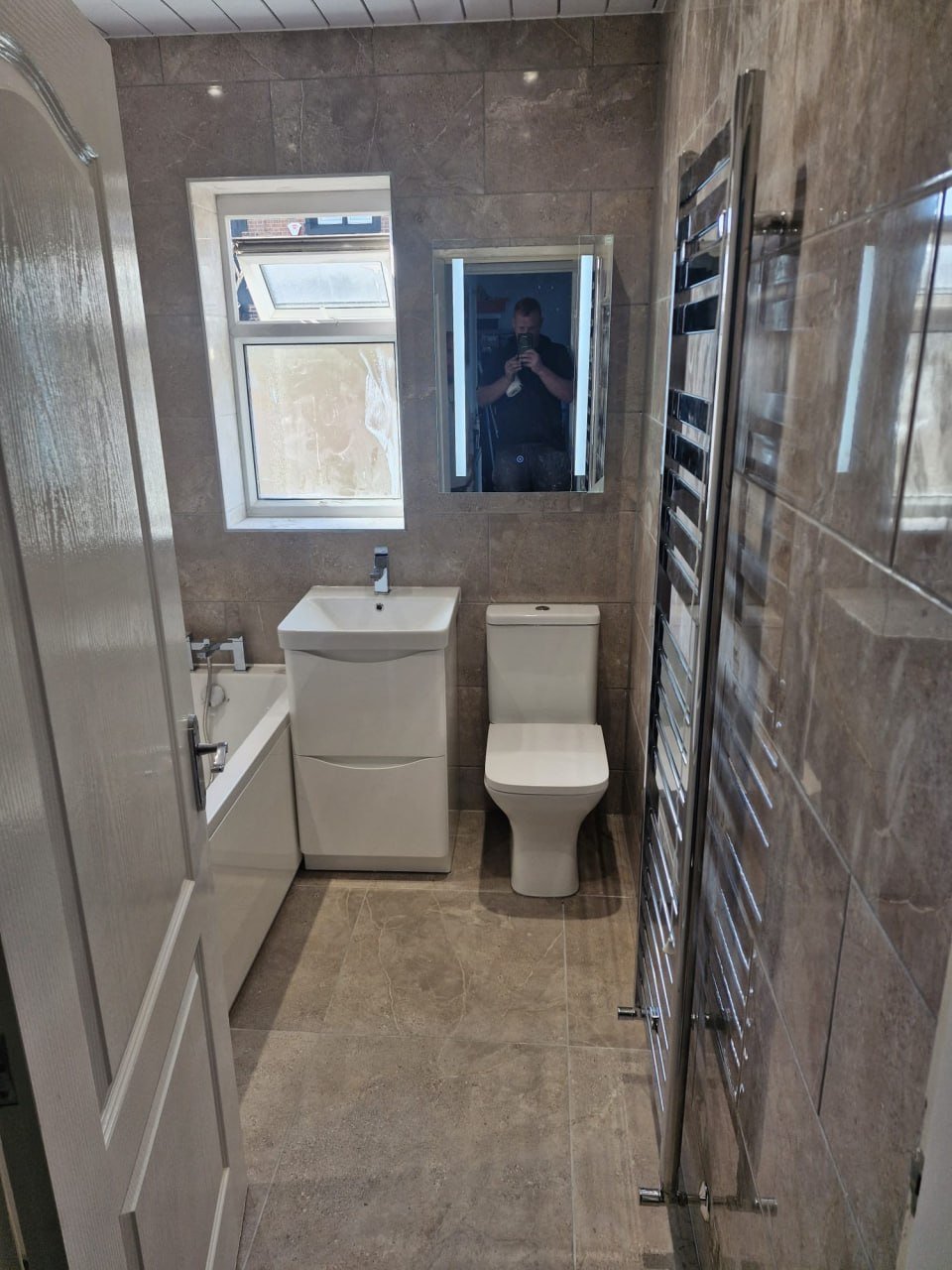 A bathroom with a white freestanding bath, a wall-hung vanity and a toilet, tiled in large grey marble-effect tiles over a wood-look floor.
