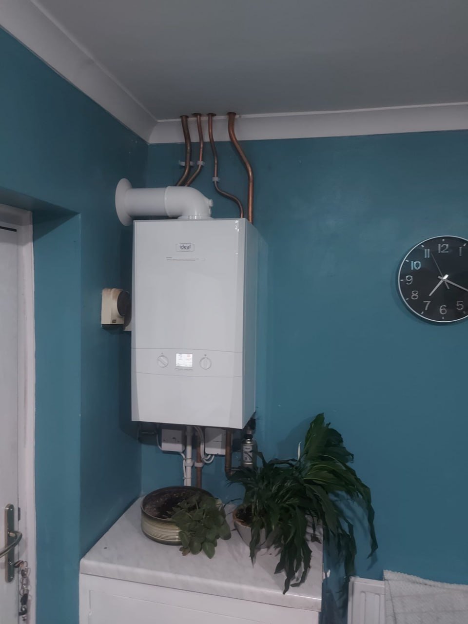 A white Ideal boiler on a teal-painted wall with copper pipes above it, a filter below and a small white control unit beside it.