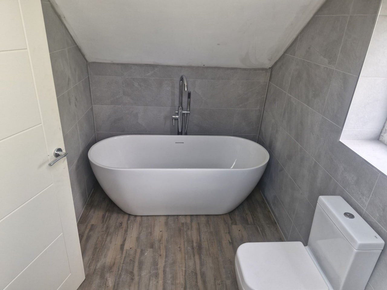 A white freestanding bath and a white toilet under a window, with large grey tiles and a wood-effect floor.