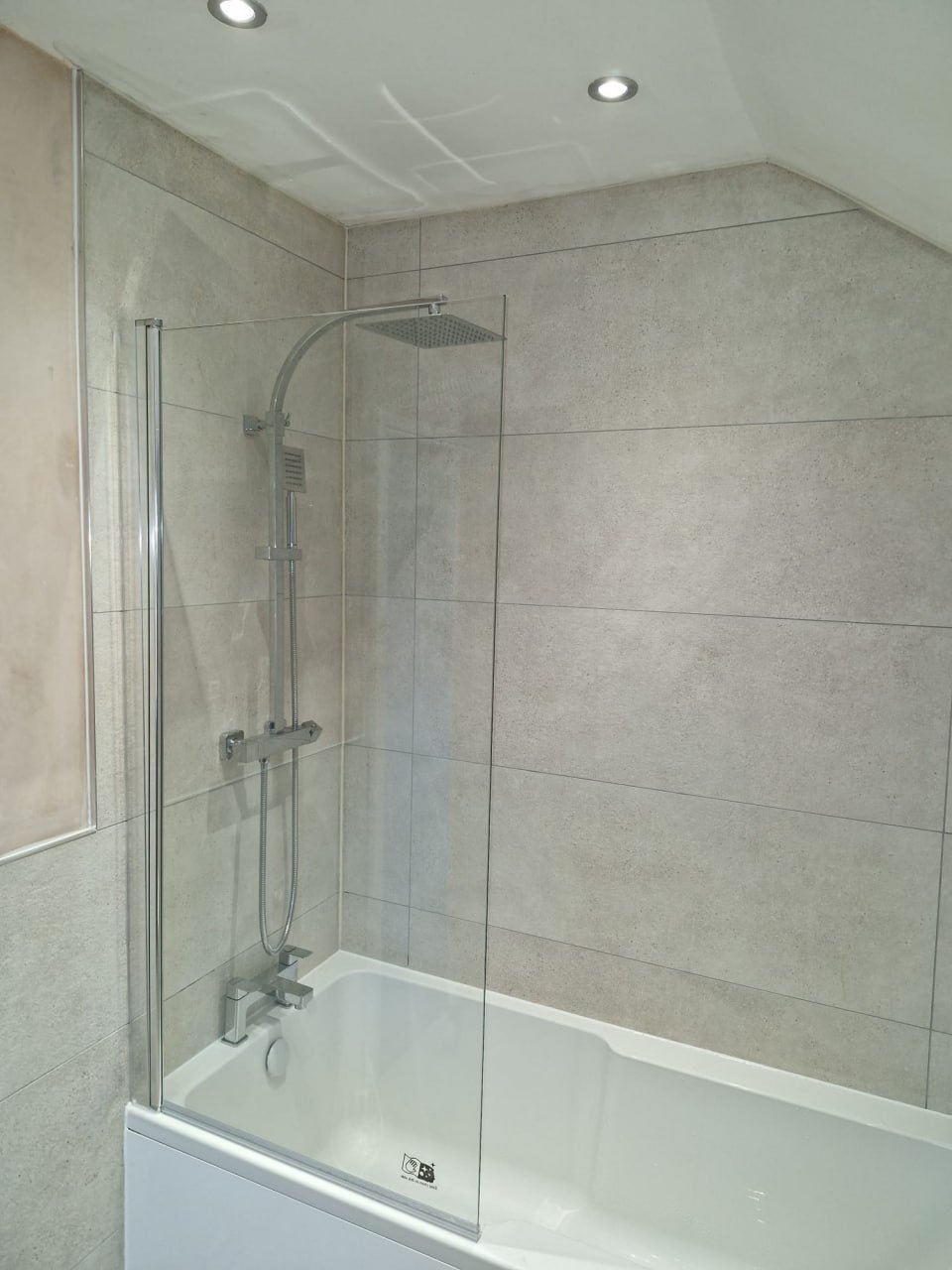 A bath with a glass shower screen, a square chrome rain shower head and a chrome mixer, against beige-grey tiled walls.