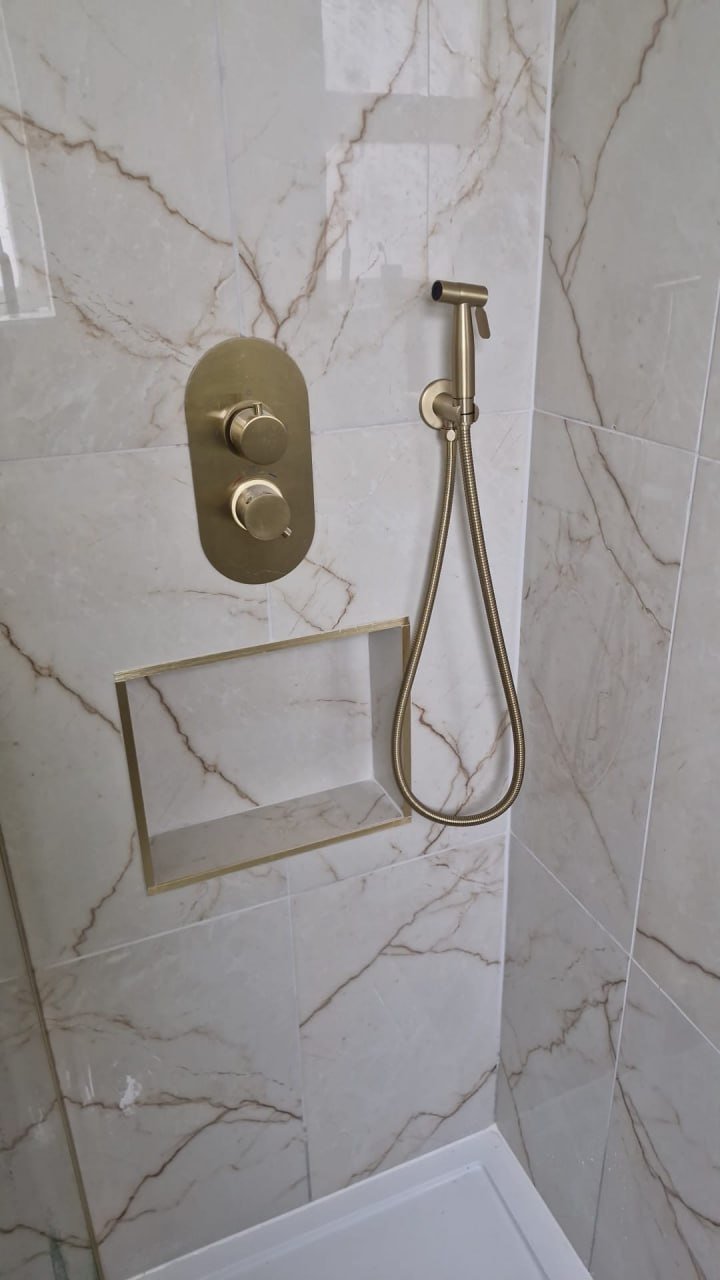 A close-up of brushed-brass shower controls and a handset on its hose, next to a recessed shelf in grey marble-effect tiling.